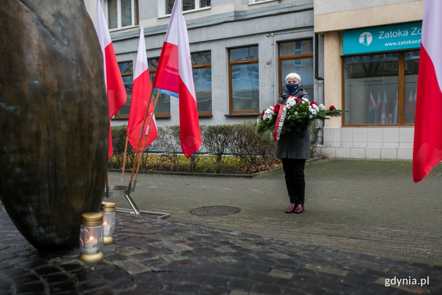 Przewodnicząca Rady Miasta Gdyni Joanna Zielińska z wieńcem w ręku stoi przed pomnikiem Ofiar Terroru Komunistycznego, przy którym palą się znicze. Przed pomnikiem, po obu stronach ustawione są biało-czerwone flagi // fot. Karol Stańczak