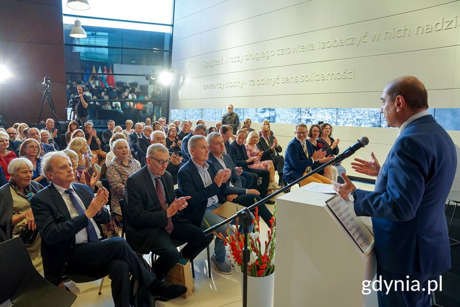 Tłum na sali, na mównicy przemawia mężczyzna.
