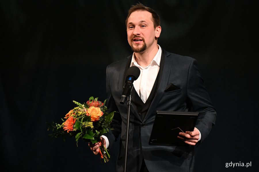 Jakub Gaweł, aktor Bałtyckiego Teatru Różnorodności, w garniturze po odebraniu Nagrody Prezydent Miasta Gdyni z okazji Międzynarodowego Dnia Teatru. W rękach trzyma kwiaty oraz dyplom 