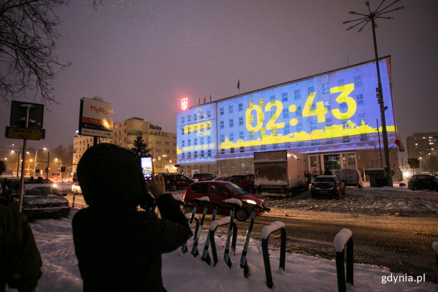 Pokaz audiowizualny wyświetlany na fasadzie budynku Urzędu Miasta Gdyni przy ul. Marsz. Piłsudskiego 52/54. Na pierwszym planie widać osobę, która robi zdjęcia budynkowi.