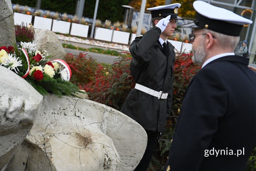 Mężczyźni w mundurach obok pomnika z szarych kamieni, na którym leżą białe i czerwone kwiaty