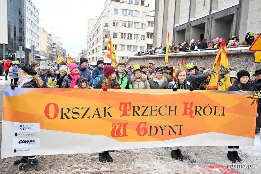 Uczestnicy gdyńskiego Orszaku Trzech Króli zgromadzeni przed kościołem pw. Najświętszego Serca Pana Jezusa z flagami w rękach i koronami na głowach. Osoby w pierwszym rzędzie trzymają baner orszaku
