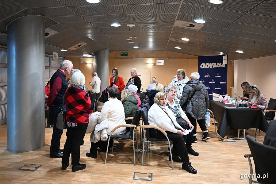 Sala konferencyjna wewnątrz hali, na środku krzesła, na których siedzą starsze osoby, po bokach stoliki