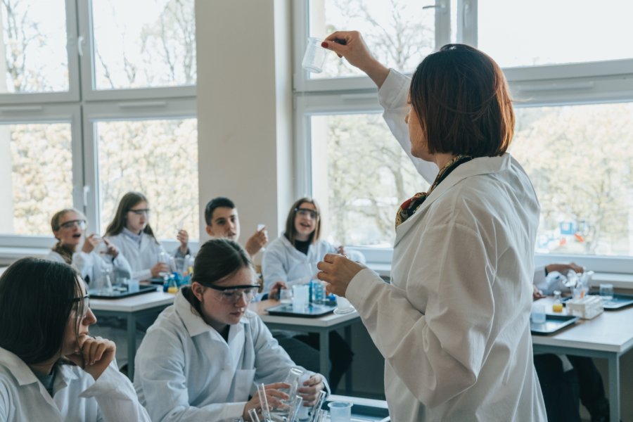 Nauczycielka w białym fartuchu, z uczniami, też w fartuchach w trakcie doświadczeń chemicznych