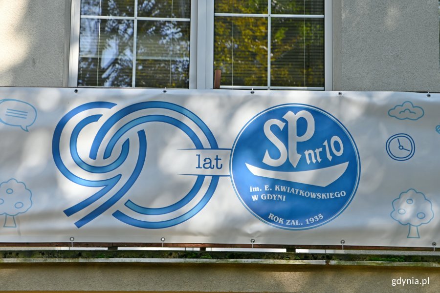 Baner na 90-lecie Szkoły Podstawowej nr 10. Na białym tle po lewej stronie zachodzące na siebie cyfry 90. Po prawej niebieskie koło z białym napisem w środku o treści: SP nr 10 im. E. Kwiatkowskiego