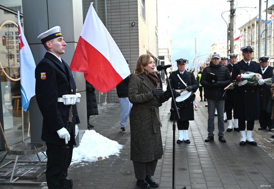 Kobieta w płaszczu przy mikrofonie, obok marynarz