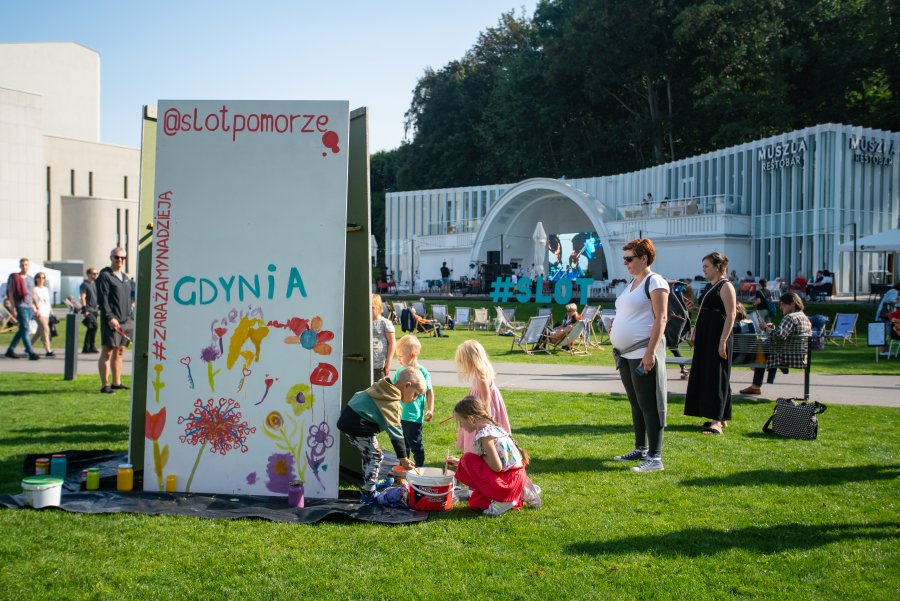 Grupa dzieci i dorosłych na placu Grunwaldzkim w Gdyni. Najmłodsi malują na białych płótnach rozstawionych na trawie. Dorośli przyglądają się ich pracy // fot. Anna Rezulak