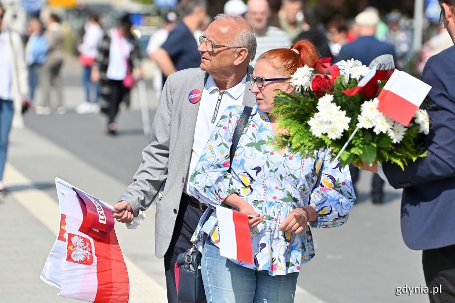 Mieszkańcy z biało-czerwonymi chorągiewkami i kwiatami na skwerze Kościuszki