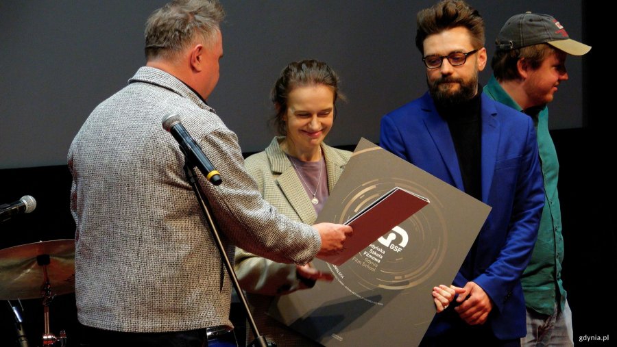 Mężczyzna w szarej marynarce wręcza kobiecie duży dyplom.