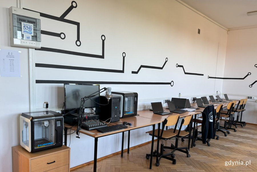 Sala ze stołami w rzędzie pod ścianą, na których stoją laptopy, w pierwszym planie drukarka 3d, dalej radiowęzeł, kolejna drukarka i laptopy