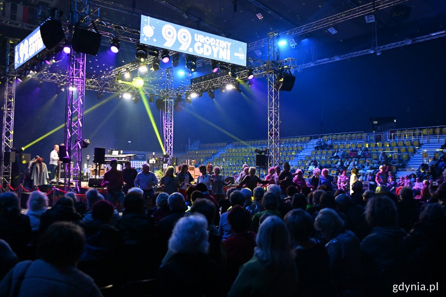 Hala Polsat Plus Arena Gdynia, scena z napisem "99. urodziny Gdyni", niebieskie światło, kolorowe oświetlenie, grupa osób stoi obok sceny