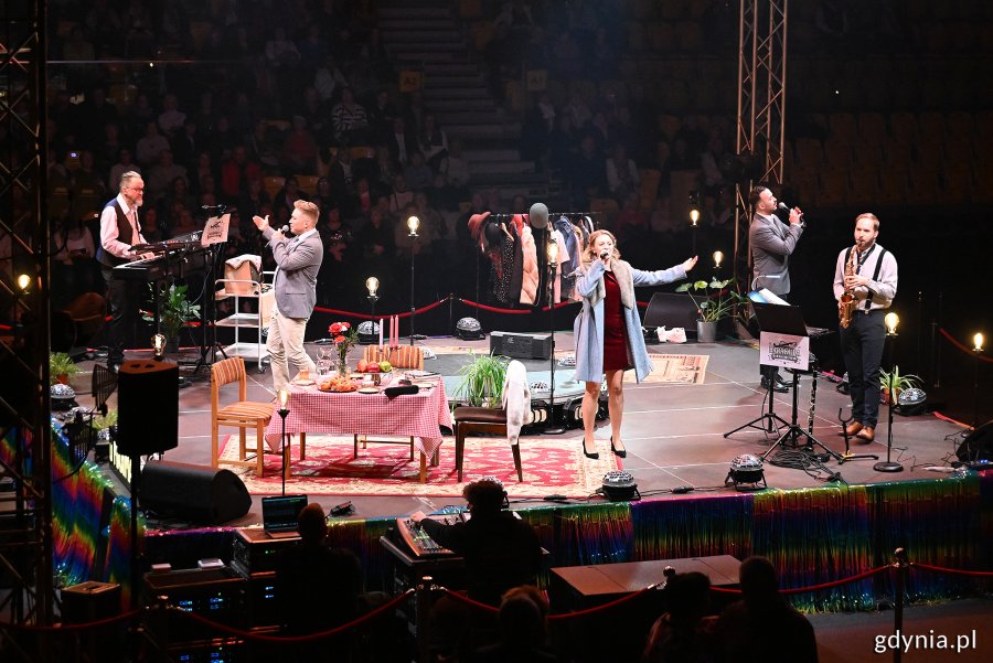 Hala Polsat Plus Arena Gdynia, scena, aktorzy na scenie, aktorka śpiewa do mikrofonu, muzycy na scenie, dekoracje, ciemność w tle