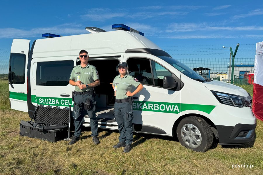 Dwie osoby ze straży celno-skarbowej na tle samochodu służbowego w kolorze białym z zielonym paskiem