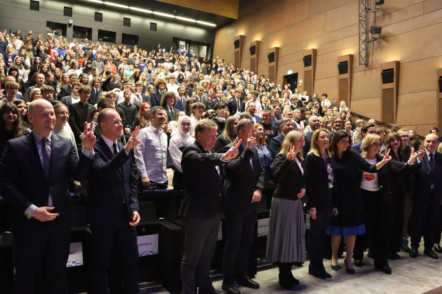 Zapełniona sala, publiczność pozująca do zdjęcia wraz z Robertą Metsola, Uśmiechnięci ludzie pokazujący do zdjęcia symbol "pokoju".