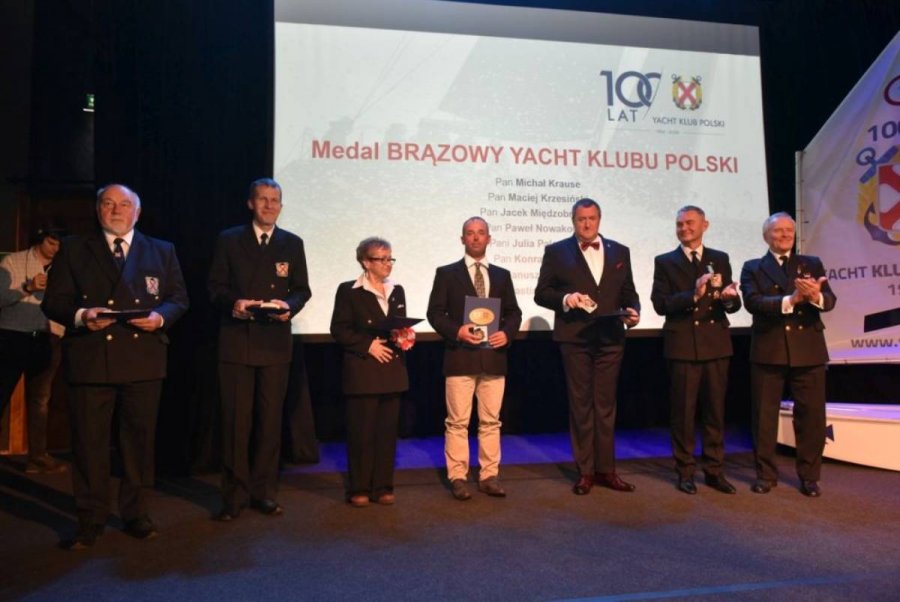 Grupa osób stojących na scenie i mających za plecami biały ekran