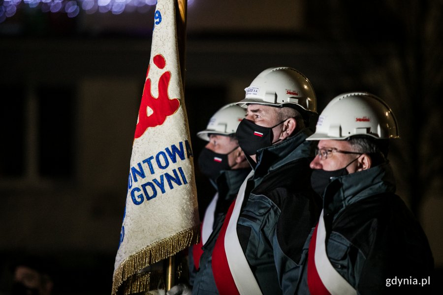 Stoczniowcy ubrani w czarne kurtki, z przewieszonymi biało-czerwonymi szarfami, w białych kaskach z logiem Solidarności podczas wieczornych uroczystości 50. rocznicy wydarzeń Grudnia '70. Jeden z nich trzyma sztandar  // fot. Karol Stańczak