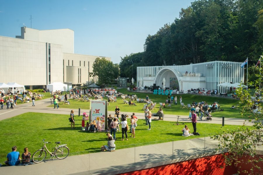 Przestrzeń miejska wypełniona ludźmi, w tle widoczne budynki teatru i muszli koncertowej. 