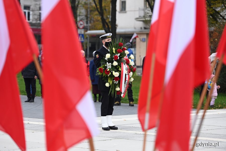 Na pierwszym planie biało-czerwone flagi. W tle marynarz trzymający w rękach wieniec // fot. Michał Puszczewicz