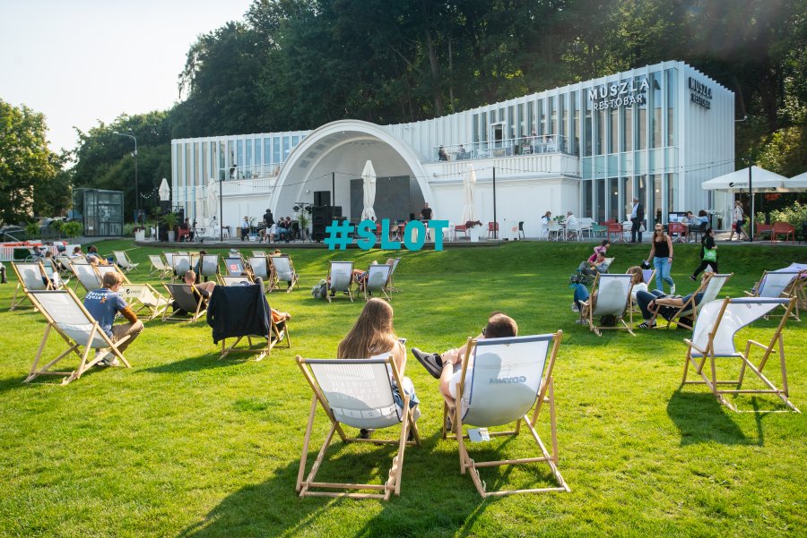 Na leżakach ustawionych na trawie siedzą uczestnicy festiwalu SLOT Pomorze. Leżaki są ustawione przed muszlą koncertową na placu Grunwaldzkim. W tle niebieski napis SLOT // fot. Anna Rezulak