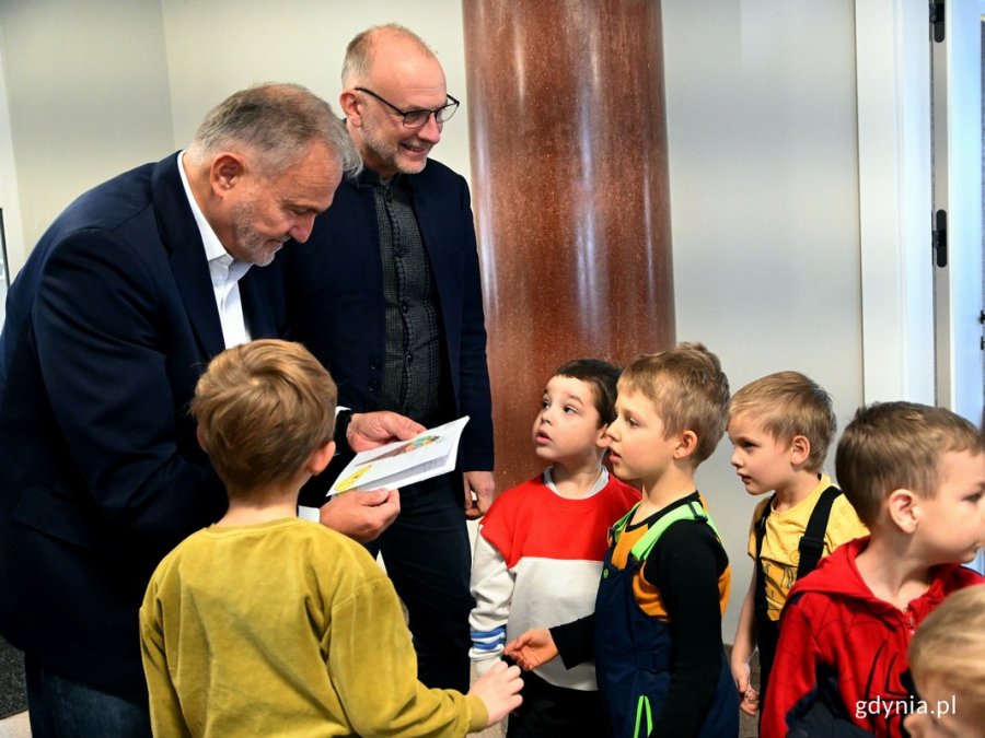 Zdjęcie przedstawia prezydenta Gdyni Wojciecha Szczurka i wiceprezydenta Gdyni Michała Gucia wraz z dziećmi wspólnie oglądają książkę z życzeniami
