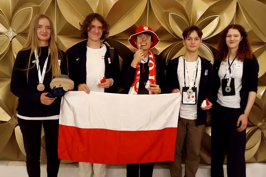 Uśmiechnieta grypa młodzieży z flagą polski przed sobą i madalami w ręka, jedna osoba ma medal w zębach, w tle złota elewacja.