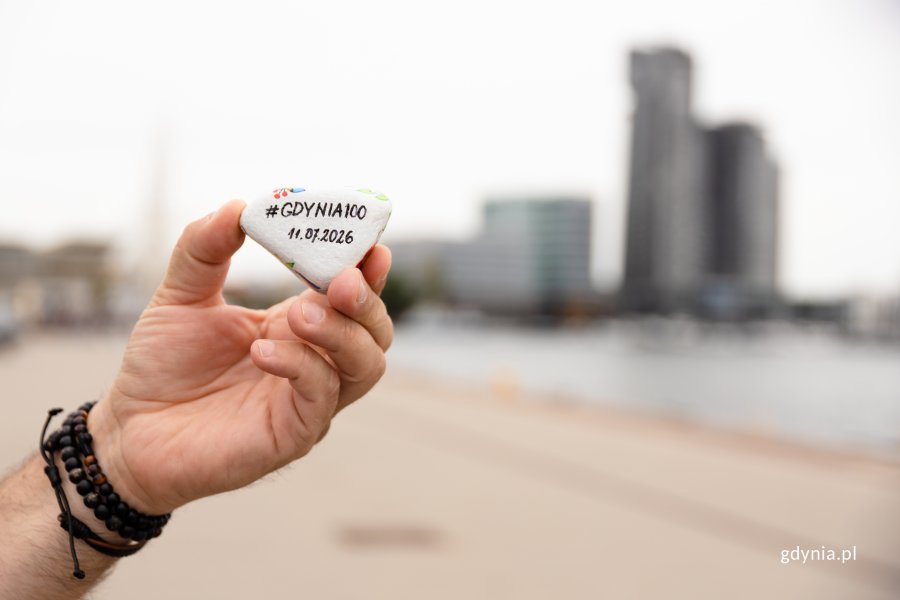 Męska dłoń na tle Sea Towers i plaży trzyma w ręku kamień z napisem #Gdynia100 i datą 11.07.2026 na 