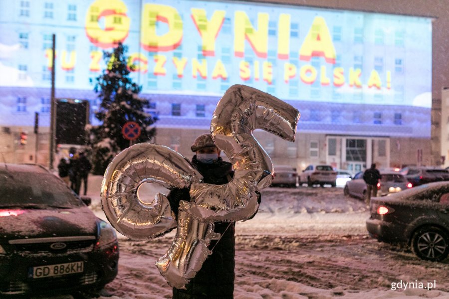 Kobieta trzymająca dwa srebrne balony wypełnione helem. Jeden balon ma kształ cyfry "9", drugi "5". W tle widoczny jest pokaz audiowizualny  wyświetlany na fasadzie budynku Urzędu Miasta Gdyni przy ul. Marsz. Piłsudskiego 52/54. // fot. Karol Stańczak