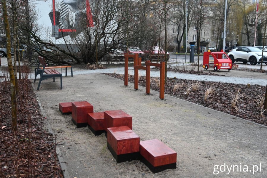 Urządzenie zabawowe w kształcie wozu strażackiego i skrzynie pełniące funkcję ławek widoczne pomiędzy ścieżkami parku