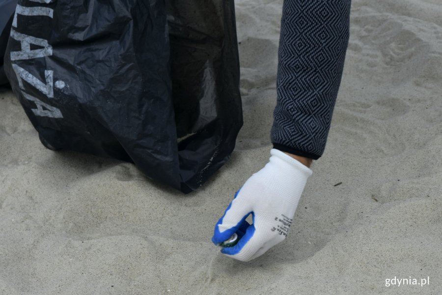 Ręka jednego z uczestników akcji - w biało-niebieskiej rękawiczce podnosi z piasku kapsel. Obok leży czarny worek na śmieci z napisem: Plaża // fot. Magdalena Czernek