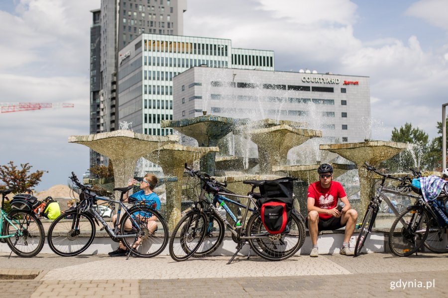 na zdjęciu rowerzyści siedzący przy fontannie w pogodny dzień, w tle Sea Towers