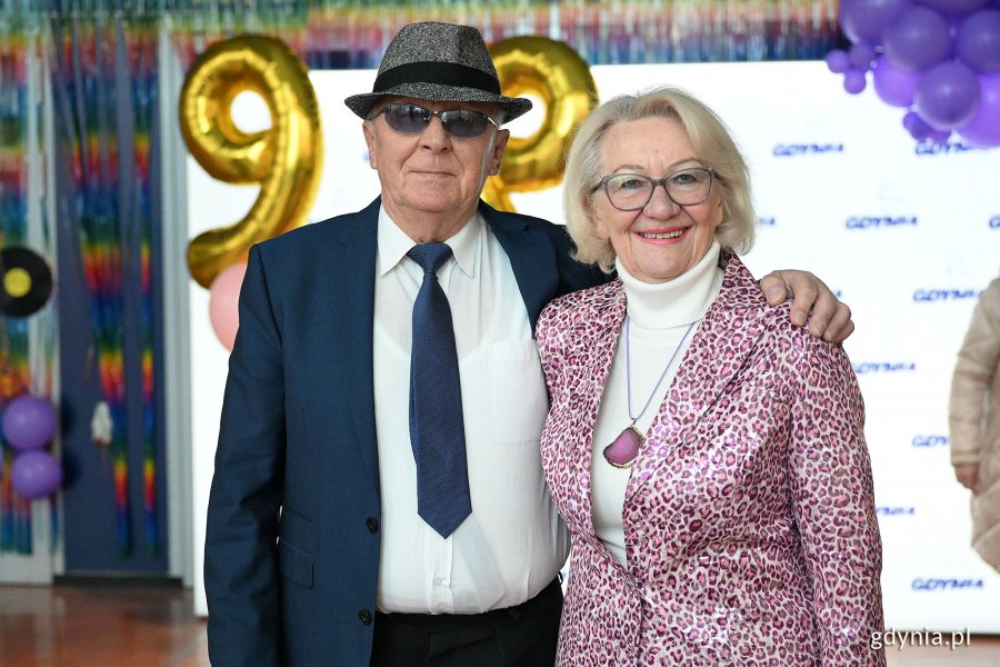 Korytarz hali, mężczyzna i kobieta w starszym wieku pozują do zdjęcia, w tle logotypy Gdyni i balony "99"