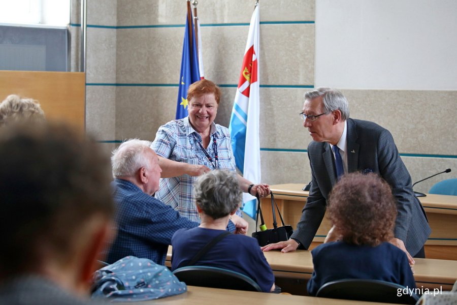 Seniorzy siedzą w sali i rozmawiają ze stojącym mężczyzną.