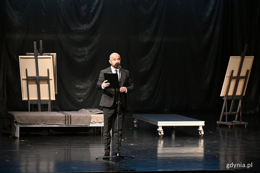Tomasz Podsiadły w garniturze, z mikrofonem i teczką w ręce podczas prowadzenia gali z okazji Międzynarodowego Dnia Teatru