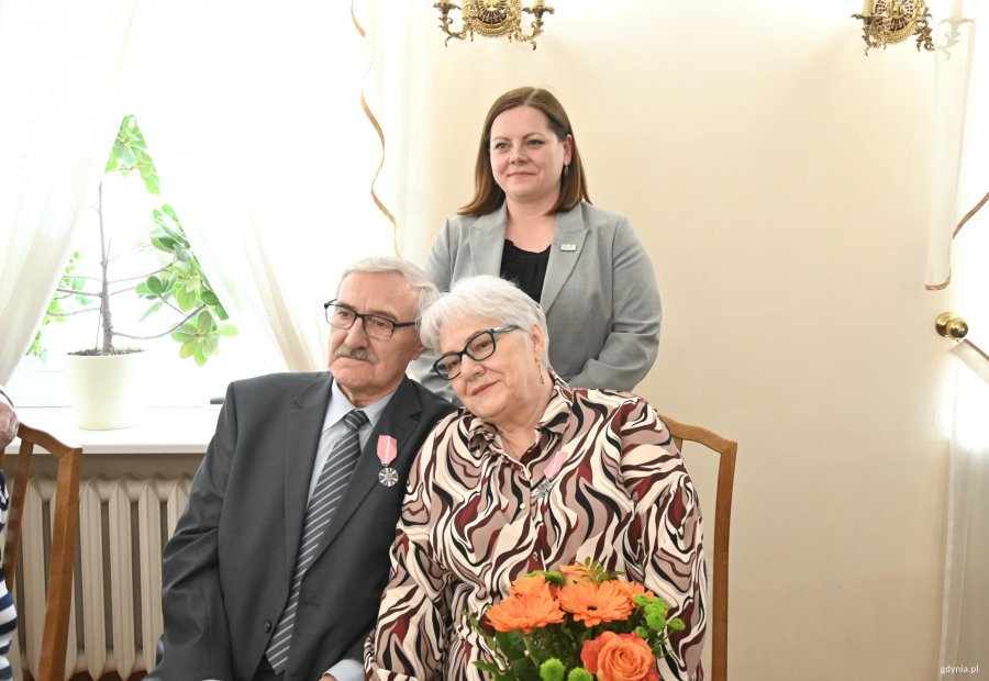 Para starszych ludzi na krzesłach. Za nimi stoi kobieta w szarej marynarce