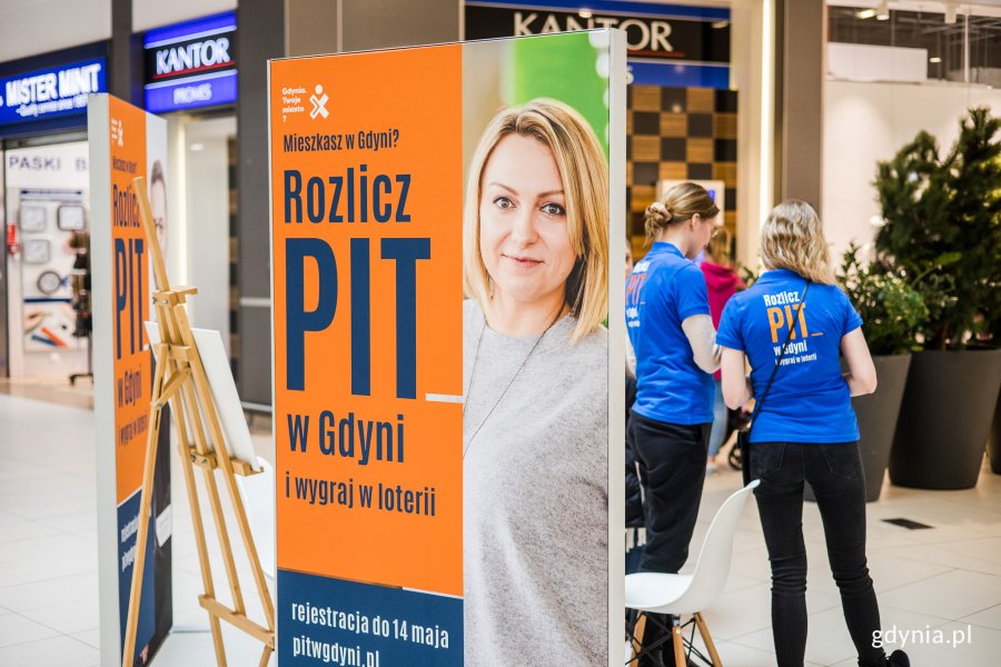 Konsultanci w Centrum Riviera przy plakacie z wizerunkiem Karoliny Kobyłeckiej-Wesołowskiej, ambasadorki tegorocznej loterii i napisem : Mieszkasz w Gdyni? Rozlicz PIT w Gdyni i wygraj w loterii