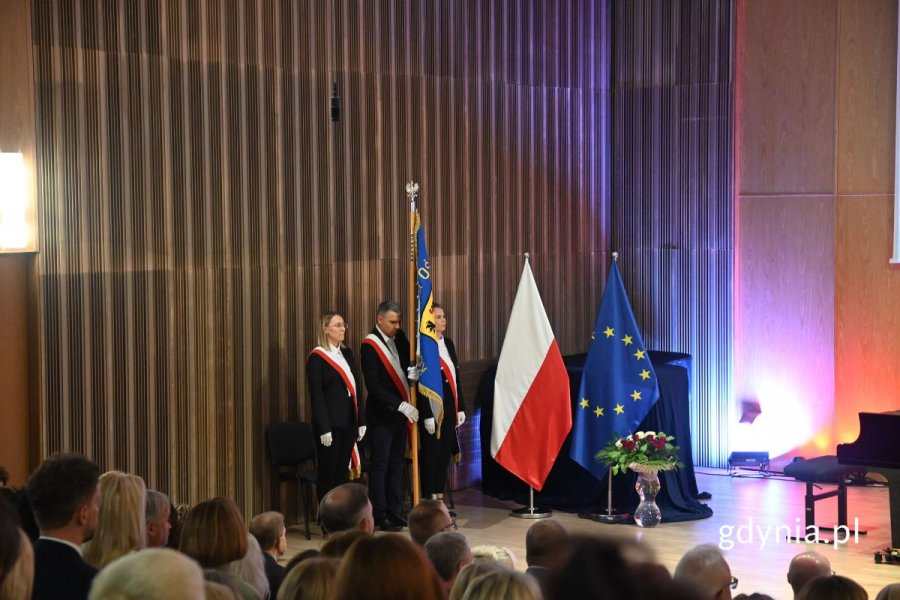 Mężczyzna i dwie kobiety stoją na scenie ze sztandarem Pomorskiego Kuratorium Oświaty. Obok flagi Polski i Unii Europejskiej