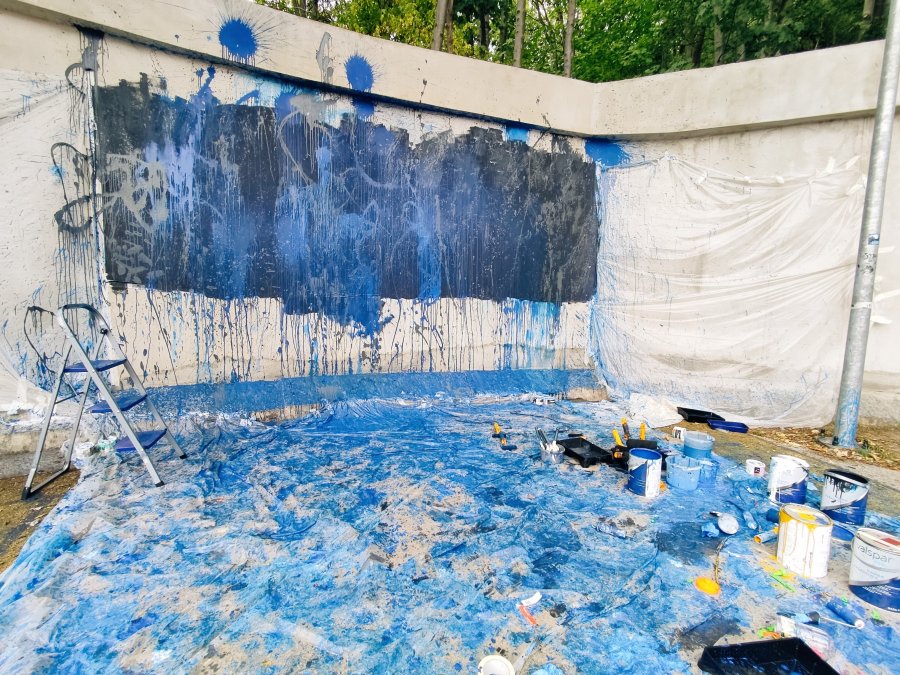 Mur, na którym namalowano mural przed wykonaniem szkicu. Na pierwszym planie rozłożona pochlapana niebieską farbą folia