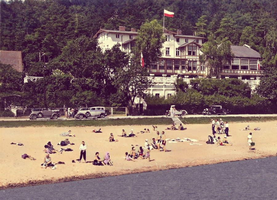 Plaża widziana od strony morza, na niej ludzie, w tle budynek i frzewa.