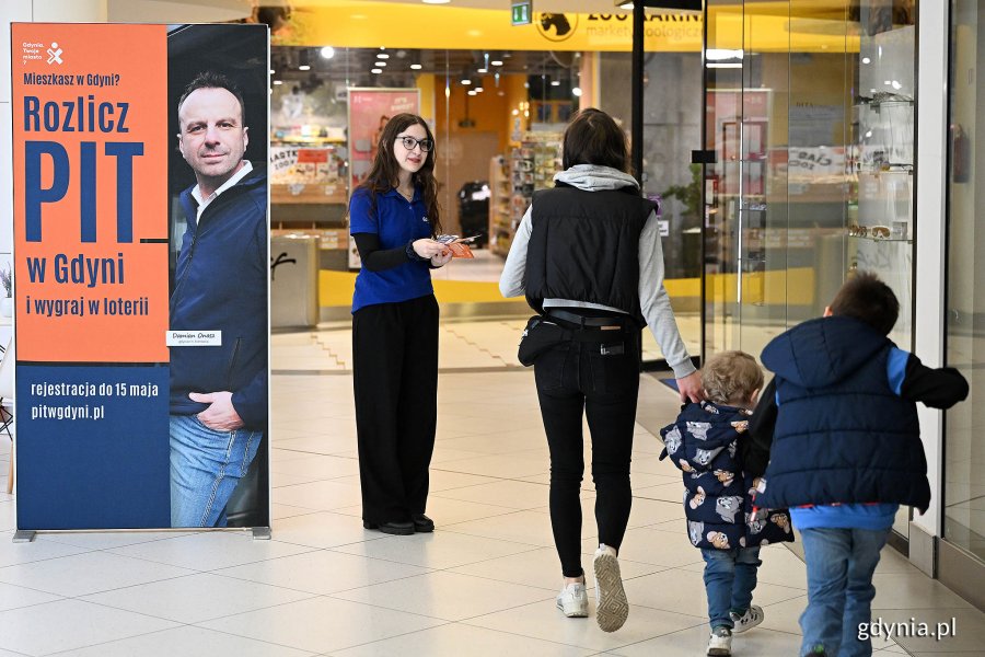 Galeria handlowa „Klif”: baner z napisem Rozlicz PIT w Gdyni i wygraj w loterii, obok konsultantka wręczająca ulotki kobiecie z dziećmi