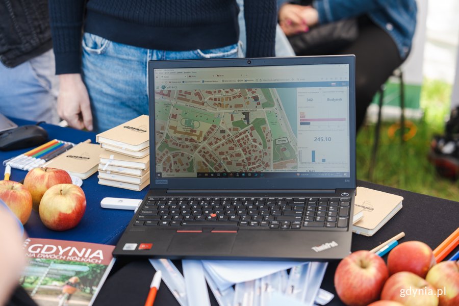 Na stoliku stoi otwarty laptop ze zdjęciem mapy Gdyni, leżą jabłka i notesy z napisem Gdynia