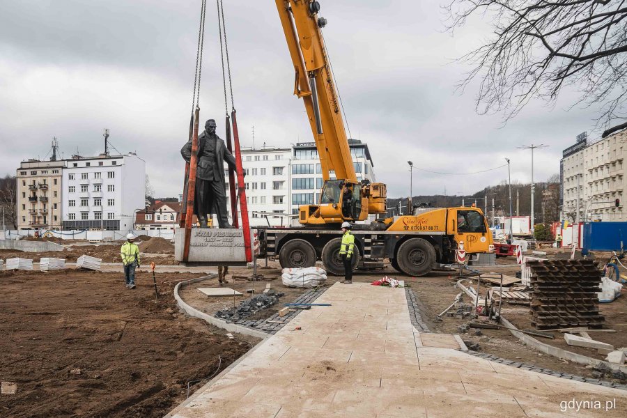 Dźwig stawia uniesiony na czerwonych taśmach pomnik Piłsudskiego w jego właściwej lokalizacji - przy skwerze Plymouth i obok budynku Urzędu Miasta Gdyni. Nad wszystkim czuwają pracownicy budowy w kaskach i żółtych kamizelkach // fot. Kamil Złoch