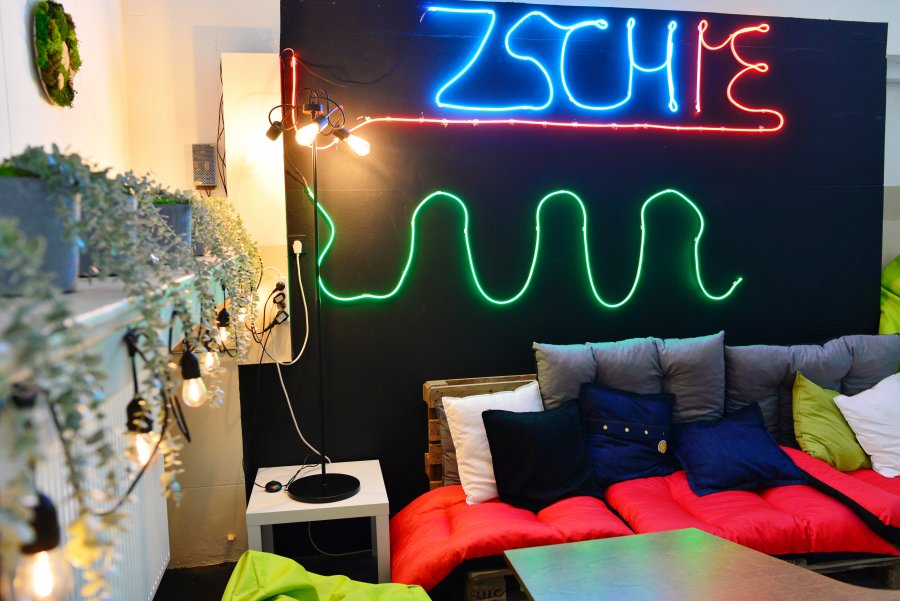 Kanapy z kolorowymi poduchami, na ścianie neonowy napis ZSChiE, na czarnym tle, po lewej półka z kwiatami, podświetlona lampkami