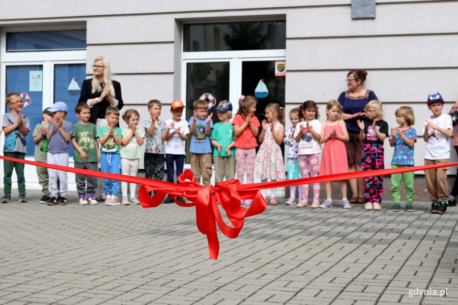 Czerwona kokarda ze wstęgą w tle dzieci stojące na tarasie przedszkola