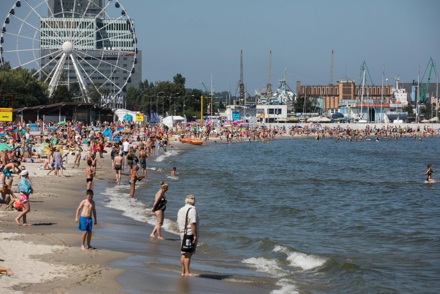 Panorama. Plaża i morze oraz wypoczywający ludzie. W tle widoczne atrakcje parku rozrywki oraz część mariny. 