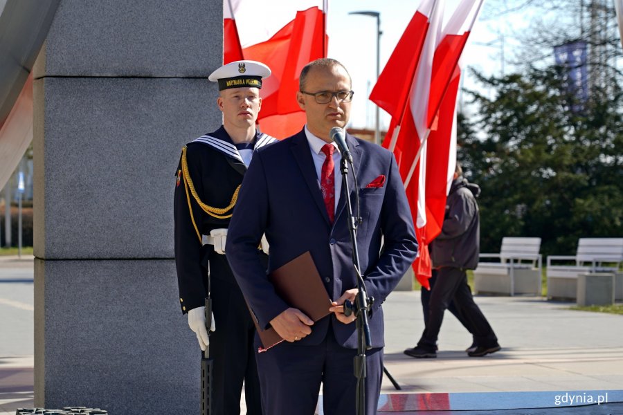 Wicewojewoda pomorski Emil Rojek przemawiający podczas uroczystości z okazji 80. rocznicy zakończenia walk o Gdynię. Za nim: marynarz z posterunku honorowego pod pomnikiem Polski Morskiej oraz powiewające na wietrze biało-czerwone flagi