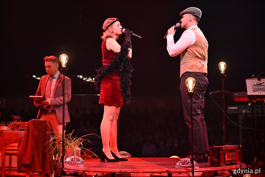 Hala Polsat Plus Arena Gdynia, scena, aktorzy na scenie, aktorka i aktor śpiewają do mikrofonu, dekoracje, czerwone światło, ciemność w tle
