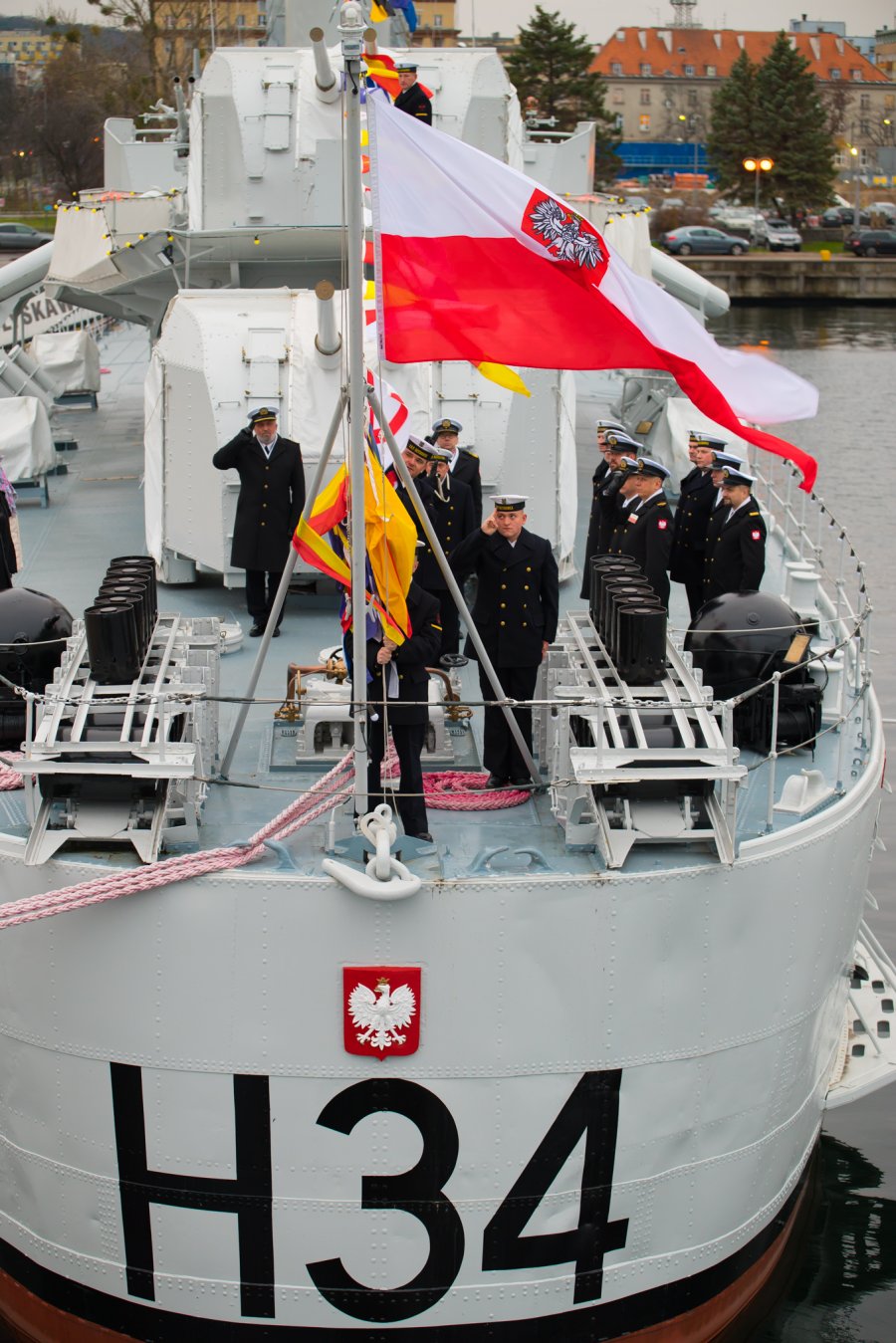 Uroczystości z okazji 84. rocznicy podniesienia bandery na okręcie ORP „Błyskawica”. Grupa marynarzy na pokładzie okrętu. Jeden z nich wciąga na maszt biało-czerwoną banderę. Pięciu stoi frontem do masztu z banderą i salutuje  // fot. bsmt Michał Pietrzak