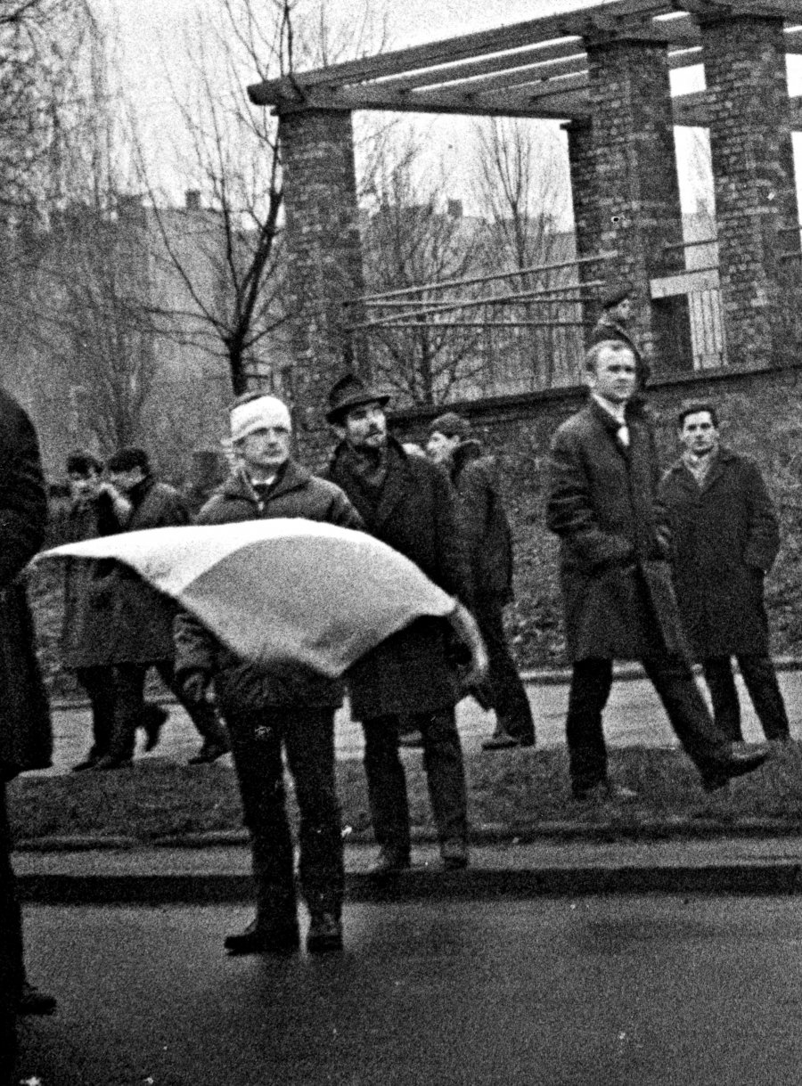 Mężczyzna z obandażowana głową trzymający flagę podczas demonstracji w grudniu 1970 r. w Gdyni na ul. Władysława IV. W głębi budynek przedszkola // źródło: IPN