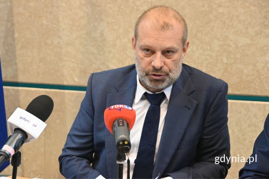 Mężczyzna w garniturze siedzący i mówiący do mikrofonu na tle beżowej ściany