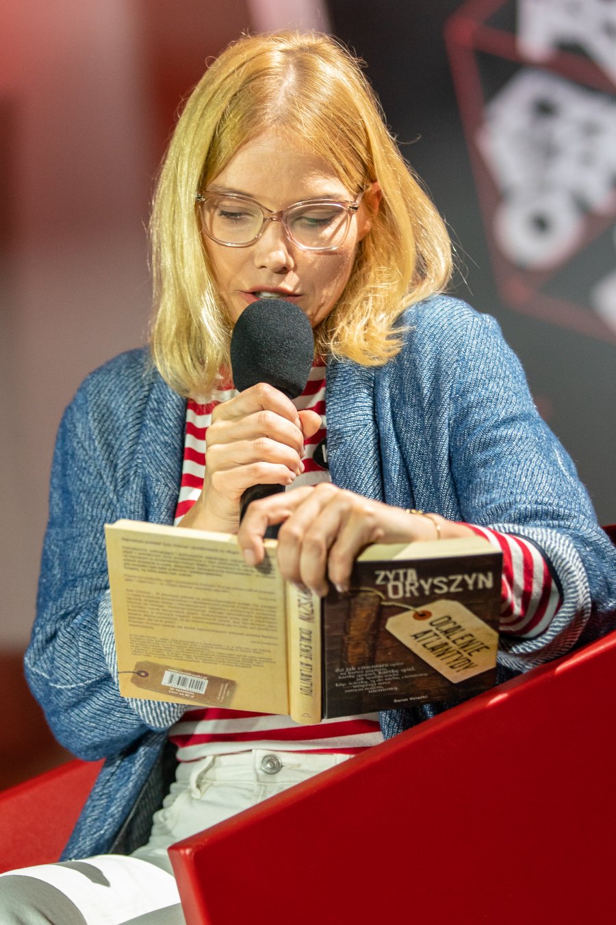 Roma Gąsiorowska - w białych spodniach, bluzce w biało-czerwone paski oraz niebieskim sweterku czyta fragment utworu Zyty Oryszyn podczas festiwalu "Miasto Słowa" w Gdyni. W jednej ręce trzyma książkę, a w drugiej mikrofon // fot. Anna Rezulak